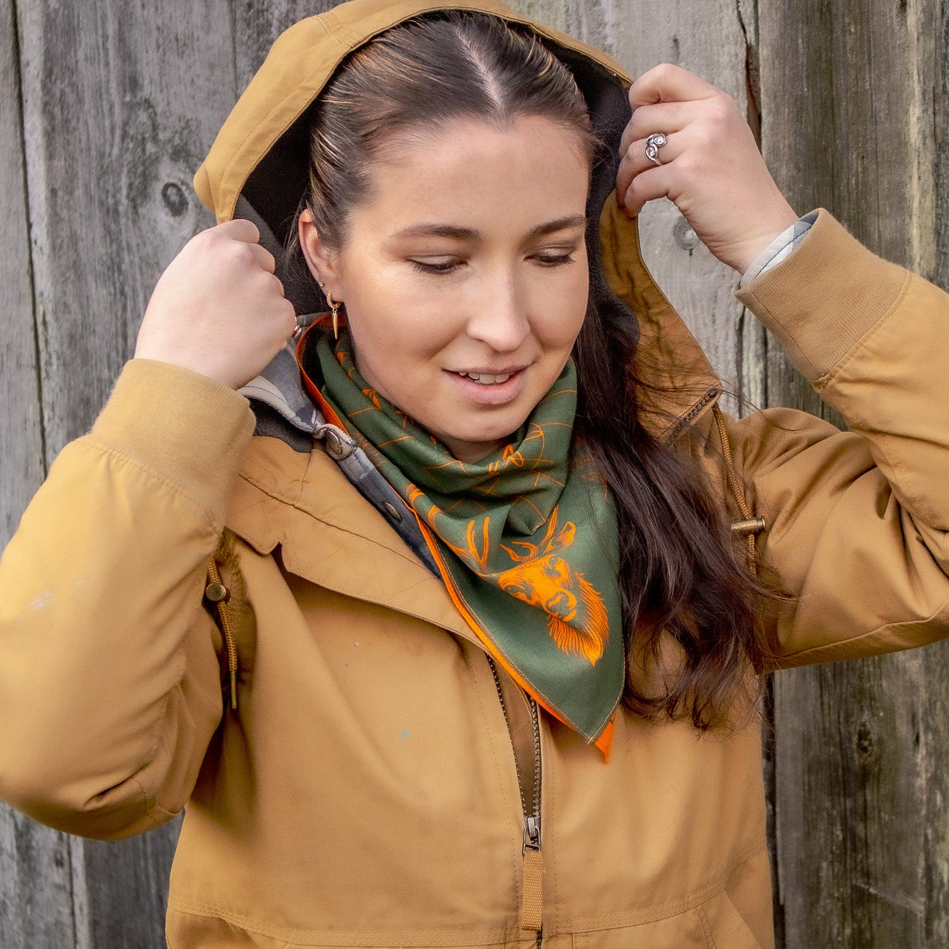 Woman wearing a mustard jacket and green scarf with orange hunter safety design against a wooden background