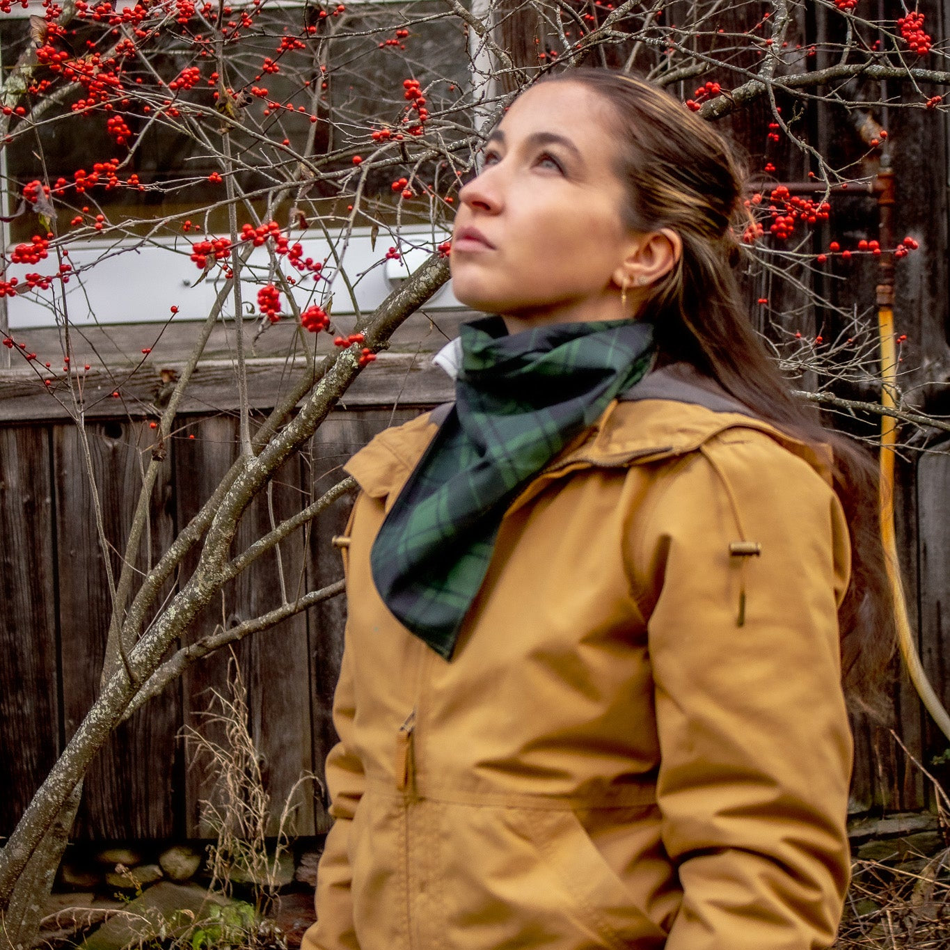 Woman in a mustard jacket and green bandana standing next to a tree with red berries bird watching