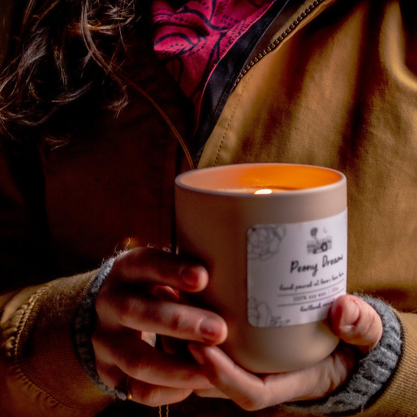 Person holding a lit candle with a label, wearing a brown jacket and gray sleeves.